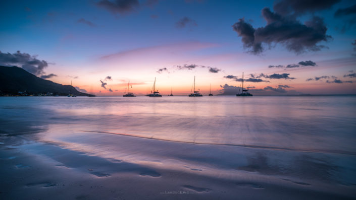 Photo of a sunset at a beach in Seychelles with boats and foot steps in sand. Buy a canvas, framed or acrylic fine art print.