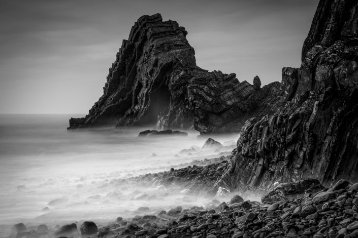 Black and white photo of rocky coast with Blackchurch Rock and waves crashing shot with long exposure in Cornwall, UK. Buy a canvas, framed or acrylic fine art print.